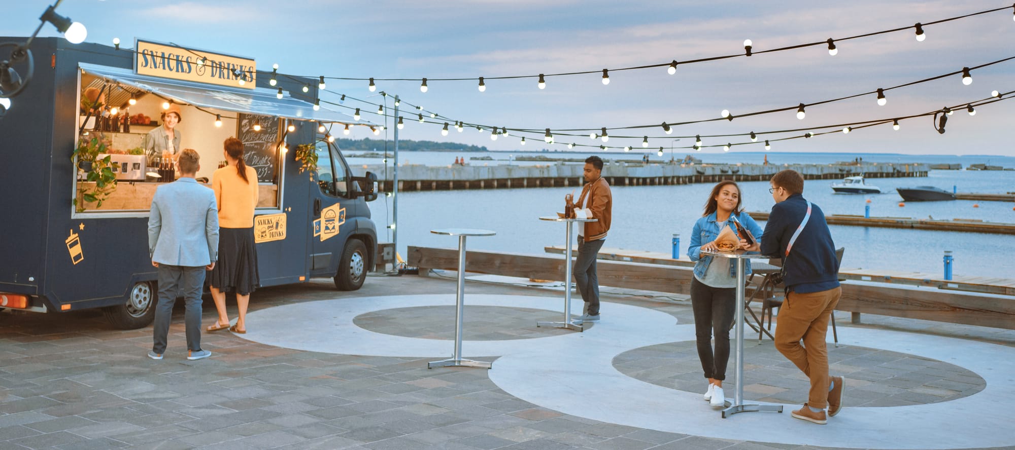 Food Truck Employee Hands Out Beef Burgers, Fries and Cold Drinks to Happy Customers. People are Eating at Tables Outside. 